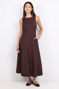 A woman modeling a sleeveless, dark brown midi dress with a high neckline and side pockets, paired with dark flat shoes, against a plain white studio background.