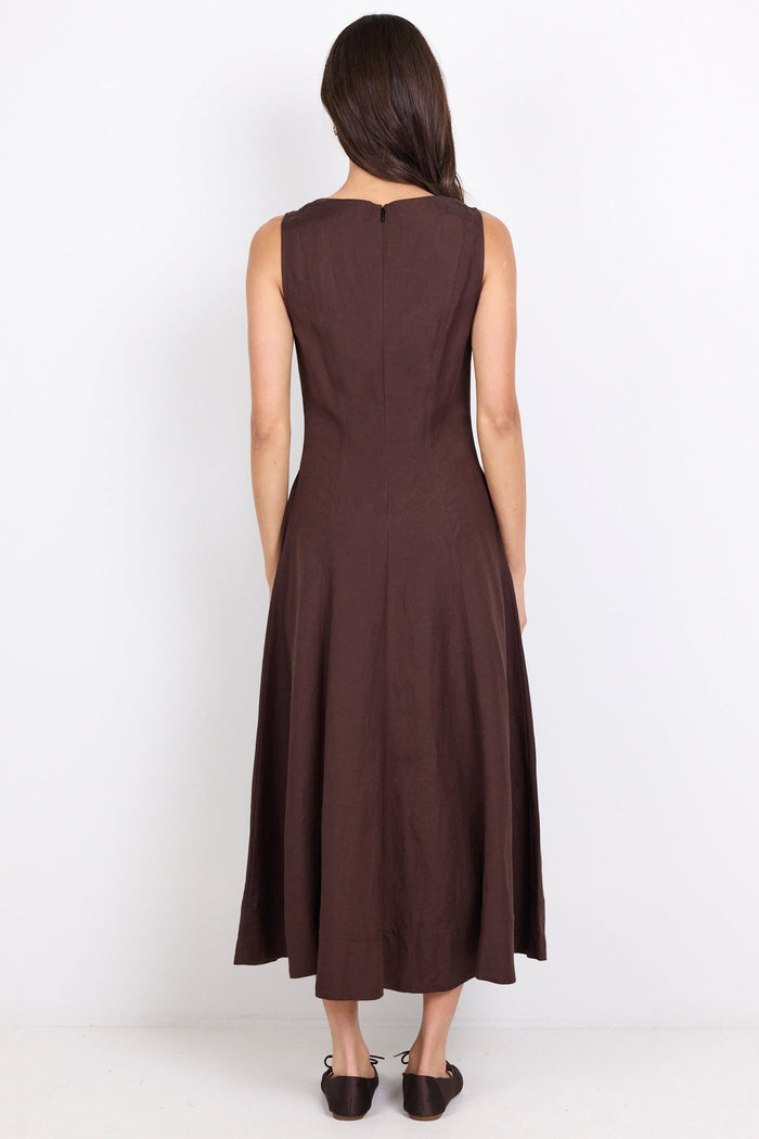 A woman viewed from the back wearing a sleeveless dark brown midi dress and black shoes, standing against a plain white studio background.