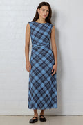 A woman stands against a white brick wall wearing a sleeveless, knee-length blue plaid dress with a gathered waist and black flats.