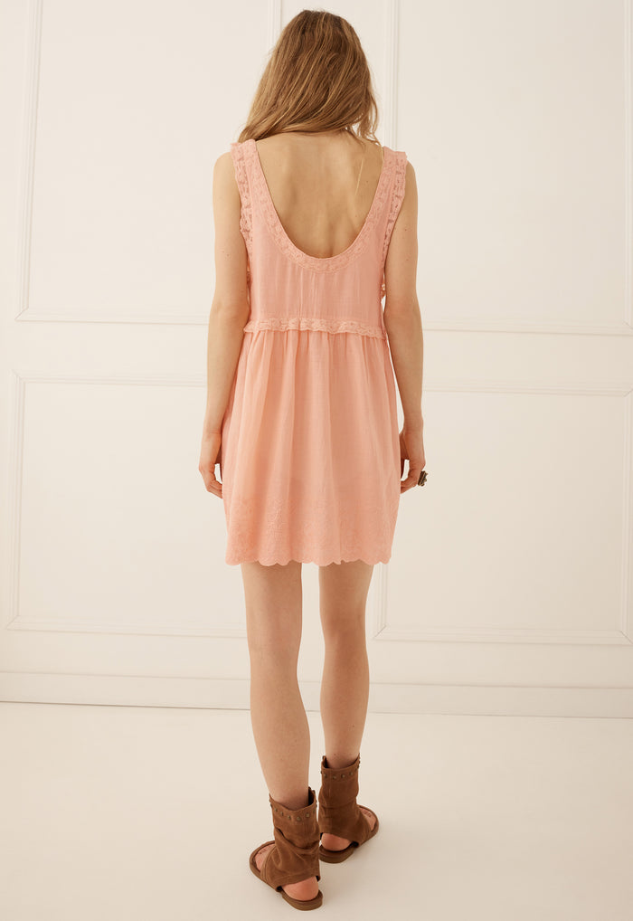 A woman viewed from behind wearing a sleeveless peach-pink mini dress with a ruffled scoop neckline and brown leather slide sandals, standing against a white paneled wall.