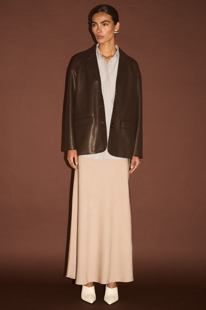 Person wearing a dark brown leather jacket over a white shirt and beige skirt on a brown background