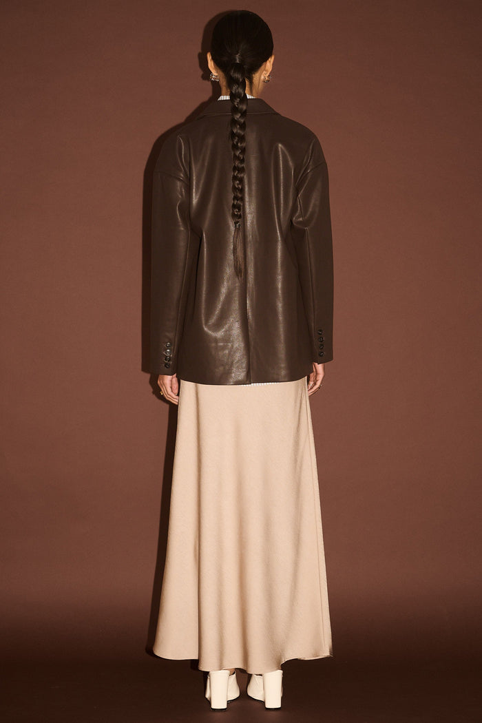 Person wearing a dark leather jacket and beige pants against a brown background