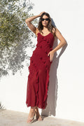 A woman wearing a long, sleeveless burgundy maxi dress with ruffled layers and sunglasses, posing outdoors against a white wall with a tree on the left.