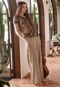 A woman stands barefoot in a rustic indoor setting with arched windows and terracotta tile flooring, wearing a bohemian-style outfit consisting of a brown patterned short-sleeve top, a wide brown belt with large silver conchos, and beige wide-leg checkered pants.