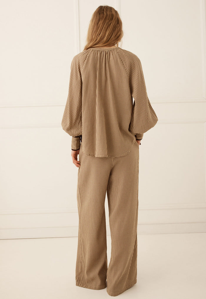 A woman viewed from the back wearing a matching beige and brown checkered two-piece set consisting of a long-sleeve blouse with balloon sleeves and wide-leg trousers, set against a clean white paneled studio background.