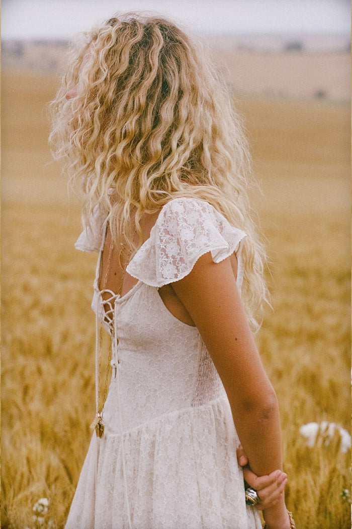 Back view of a model in Glastonbury Lace Dress, lace-up back detail and flutter sleeves, wheat field setting