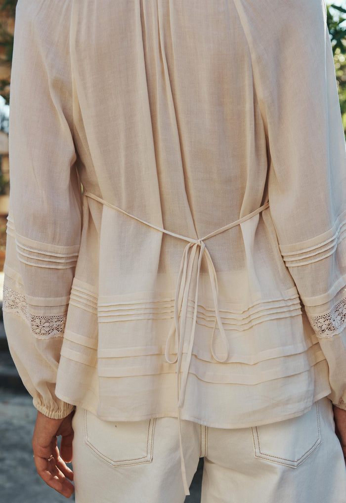 Back view of Hera Lace Blouse showing pintuck hem detail, back waist tie, and lace-trim sleeves in macadamia