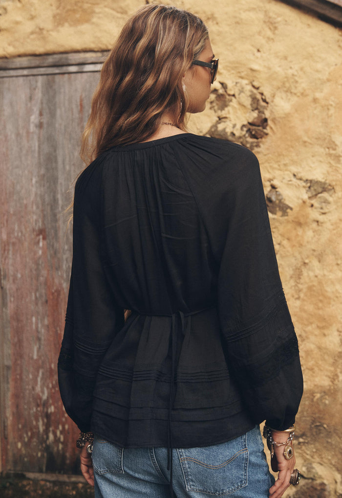 Back view of a model wearing Hera Lace Blouse in Vintage Black, showing back waist tie and pintuck hem detail