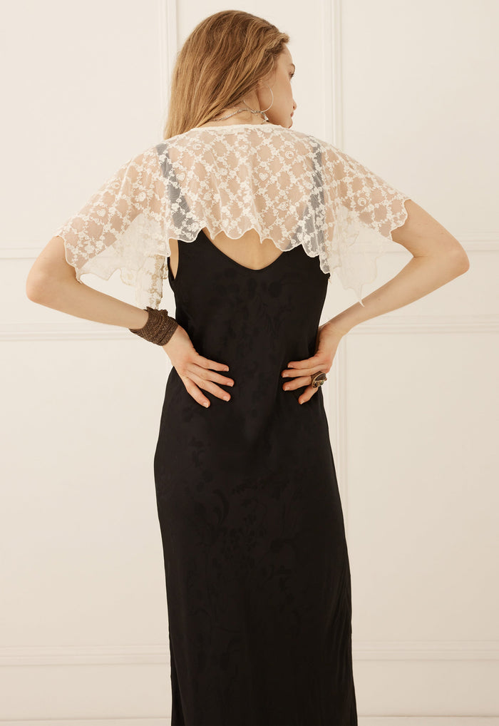 A woman shown from the back wearing a black maxi dress with a sheer white lace cape-style shoulder overlay, standing against a white paneled wall.