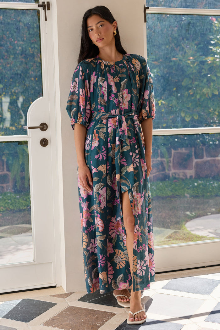 A woman models a floral midi dress with a high slit, standing in a doorway with a garden view in the background. The dress features a dark teal base with pink, purple, and green tropical floral patterns.
