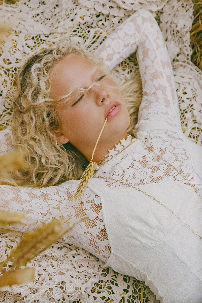 Model lying down, cream lace top with long sleeves and high neck detail visible against lace backdrop