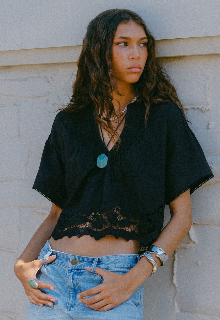 Front-facing model shot of black organic cotton-linen crop blouse with lace hem trim and V-neck tie detail