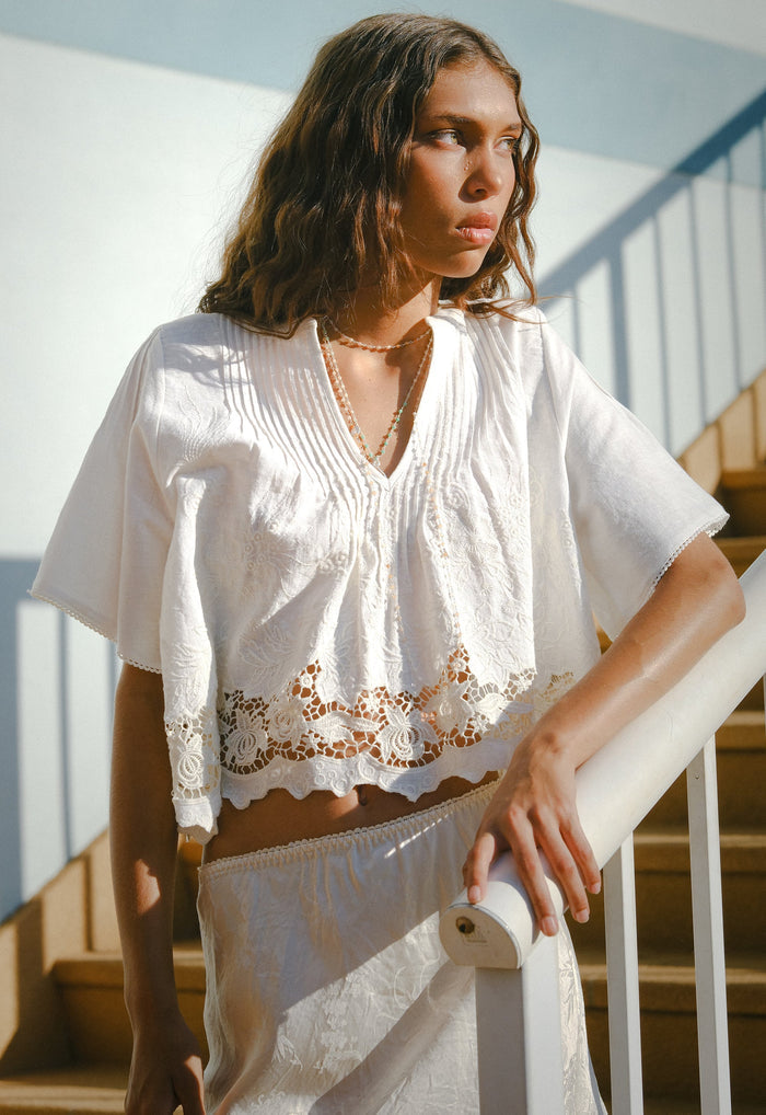 Front view of white organic cotton-linen blouse with crochet lace hem, pintuck detail, and V-neck on staircase