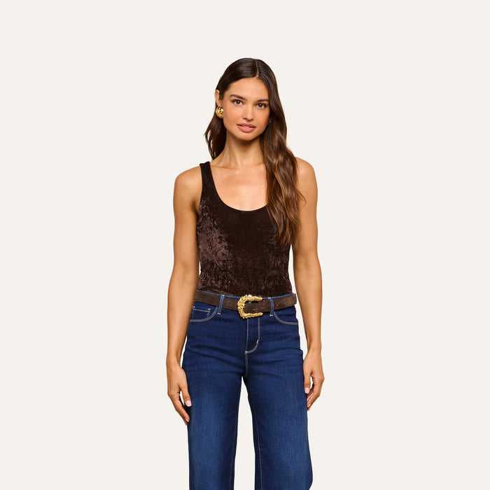 A woman with long brown hair stands against a plain off-white background, wearing the Kalea Velvet Bodysuit in dark brown, styled sleeveless and paired with blue jeans featuring a gold buckle belt and a small gold earring.