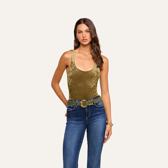 A woman stands against an off-white background wearing the Kalea Velvet Bodysuit in sleeveless olive-green velvet with a scoop neckline, paired with blue jeans, a wide tan belt, and a bracelet.