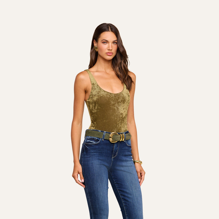 A woman with long, wavy brown hair and a neutral expression is standing against a plain background, wearing the Kalea Velvet Bodysuit in olive green featuring a scoop neckline. She pairs it with blue jeans, a wide belt, and a bracelet on her right wrist.