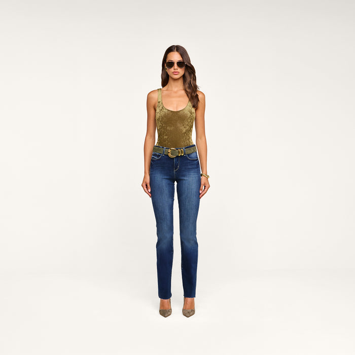 A woman wearing sunglasses, the Kalea Velvet Bodysuit in sleeveless gold velvet with a scoop neckline, blue jeans with a decorative belt, and beige heels stands against a plain white background.