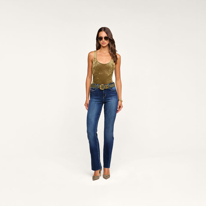 Woman wearing sunglasses, the Kalea Velvet Bodysuit with a scoop neckline, blue jeans with a double-ring belt, and pointed heels stands against a plain white background.