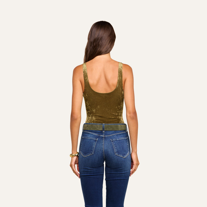 A woman with long brown hair, dressed in the Kalea Velvet Bodysuit in olive green with a scoop neckline and paired with blue jeans, stands facing away from the camera against a plain light background.
