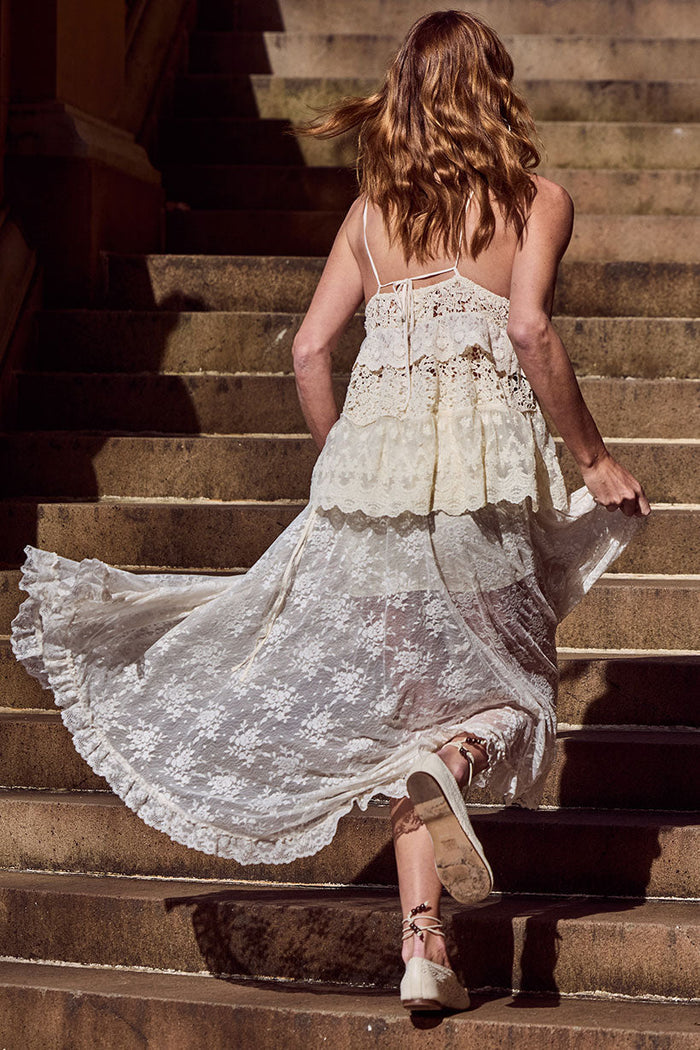 Back view of a model walking upstairs in cloud cream lace maxi skirt, showing open tie back and floral lace tiers in motion
