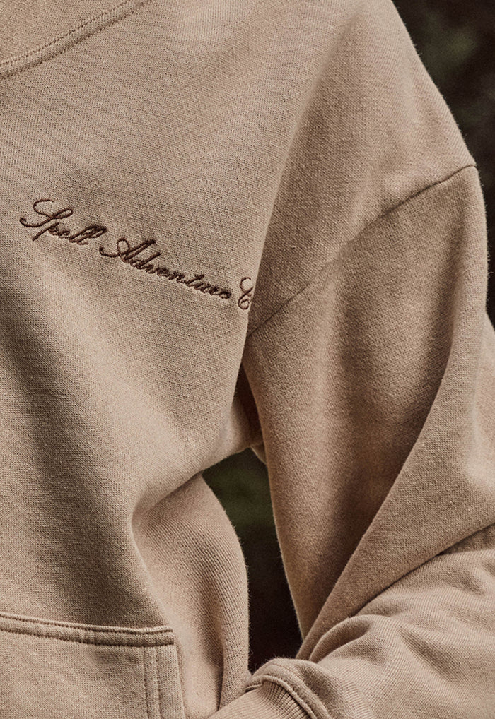 Close-up of a beige crewneck sweatshirt with dark brown embroidered script on the chest reading 'Spell Adventure Club'.