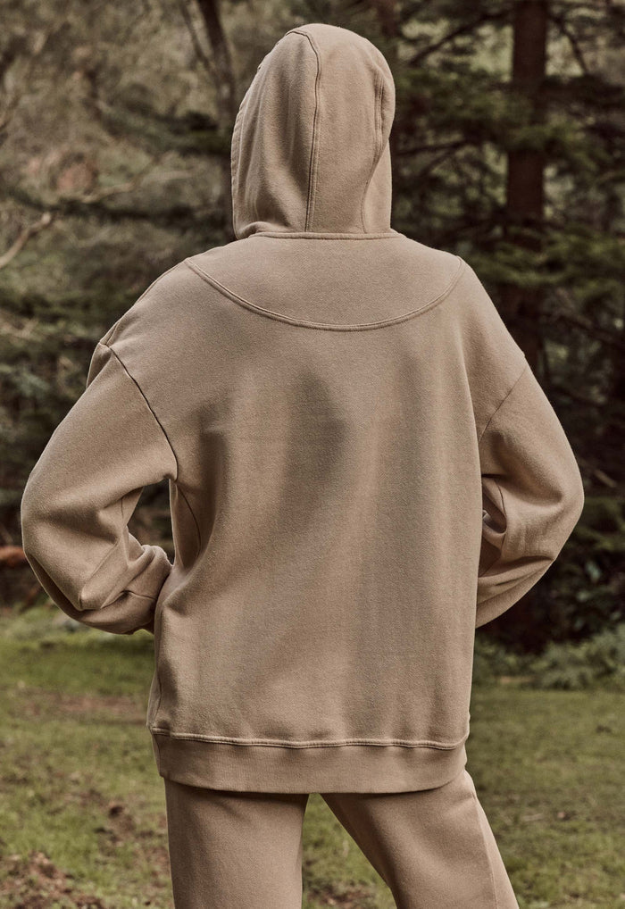A person viewed from behind wearing a matching beige hooded sweatshirt and jogger pants in an outdoor wooded setting.