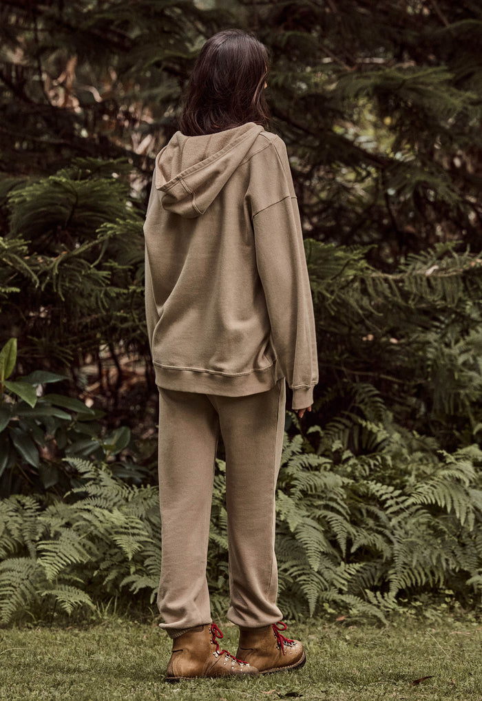 A person standing outdoors in a wooded area, viewed from behind, wearing a matching beige hooded sweatshirt and jogger pants with brown lace-up boots.