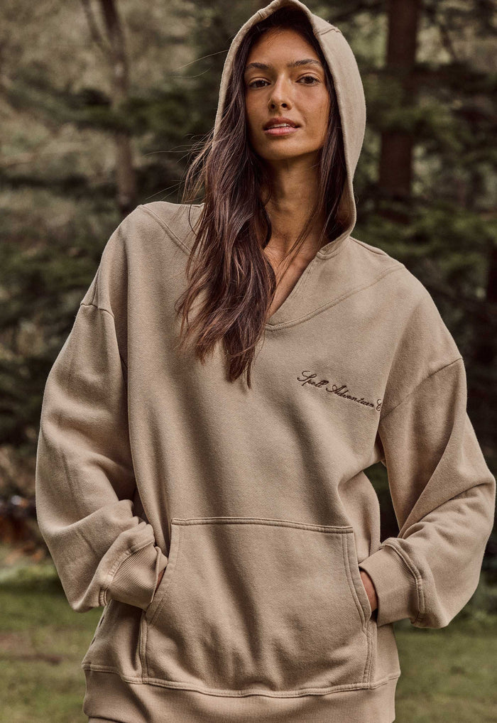 A woman wearing a beige oversized pullover hoodie with a small script logo on the left chest, photographed outdoors against a blurred forest background.