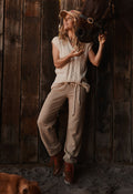A woman in a rustic barn setting wearing a cream knit sleeveless top, beige drawstring sweatpants, and brown boots, posing with a horse halter and looking upward. A dog is partially visible in the lower left corner.