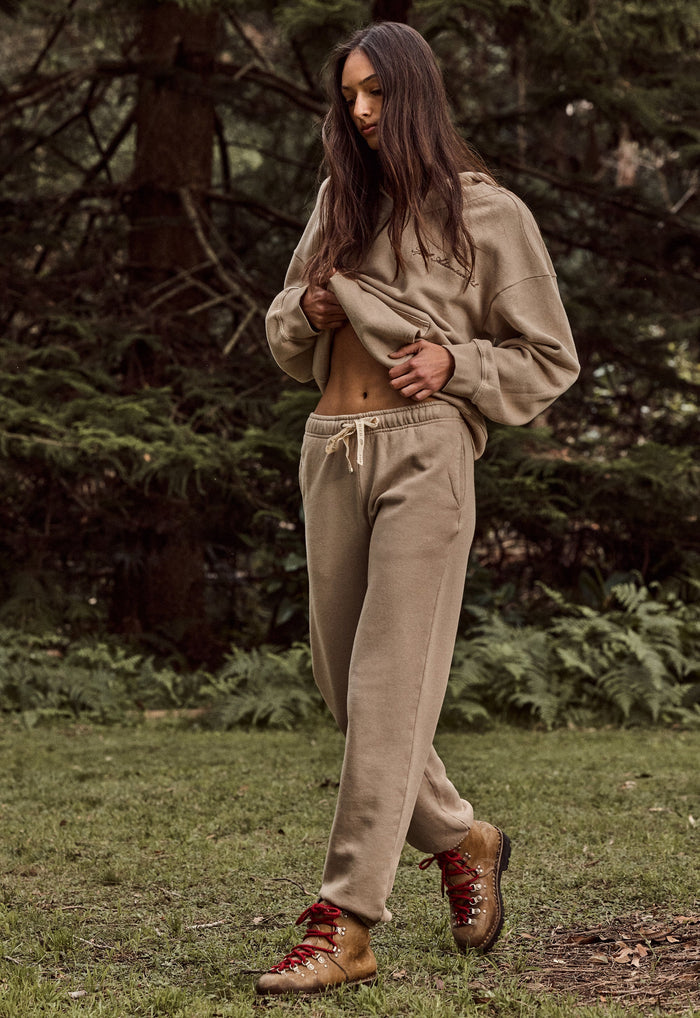 A woman wearing a matching beige cropped hoodie and sweatpants set with brown lace-up boots, standing in a grassy forest clearing.