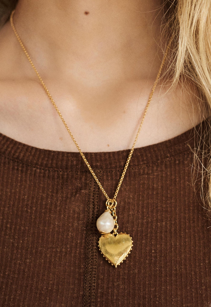Close-up of gold-plated heart locket pendant with baroque pearl charm on fine chain, worn over brown ribbed top