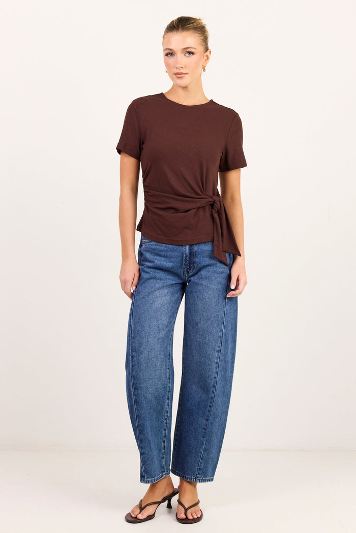 A woman modeling a dark brown short-sleeve knit top with a twisted front detail and side tie, paired with blue jeans and black sandals, against a plain white studio background.
