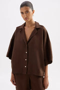 A woman wearing a dark brown short-sleeve button-up shirt and matching brown trousers, photographed against a plain light gray studio background.