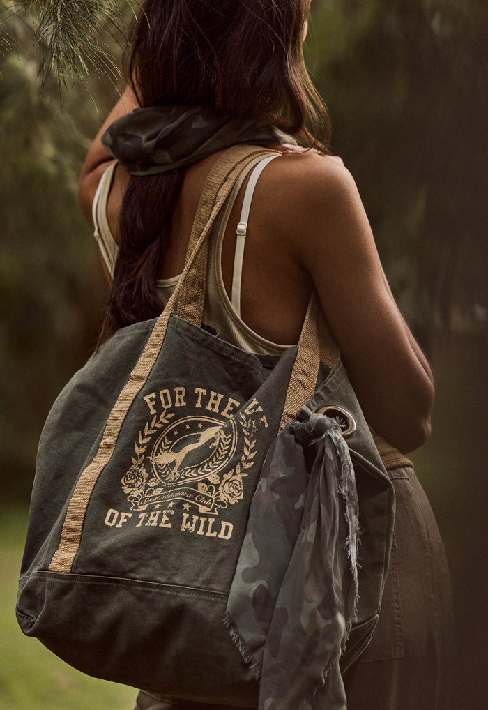 A woman outdoors carrying a large canvas tote bag over her shoulder. The bag is dark olive green with tan straps and a tan graphic print that reads 'FOR THE LOVE OF THE WILD' around a illustration.