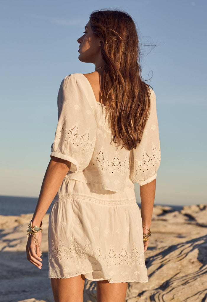 Back view of antique white organic cotton voile blouse with broderie anglaise hem and elbow sleeves, rocky beach setting
