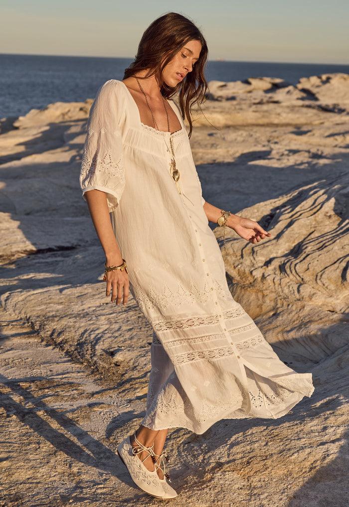 Front view of a model walking on coastal rocks in white organic cotton voile midi dress with eyelet hem and lace trim bands