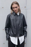 Person wearing a black leather jacket over a white shirt against a gray concrete wall.