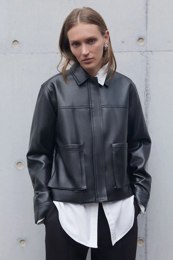 Person wearing a black leather jacket over a white shirt against a gray concrete wall.