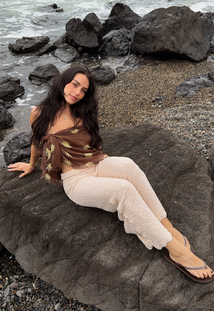 Model seated on coastal rocks wearing Melo Brown modal scarf tied as a bandeau top with gold sun print