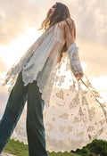 A woman outdoors wearing a sheer white lace long-sleeve cardigan with gold butterfly and moon motifs over dark teal wide-leg jeans, photographed against a bright sunset sky.