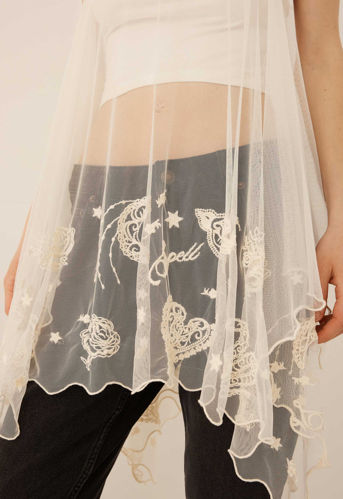 A close-up of a person wearing a sheer white lace overlay draped over dark gray jeans, with a white crop top partially visible. The lace features embroidered celestial motifs including stars, hearts, and the word 'spell' in script.