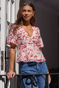 A woman wearing a short-sleeve wrap-style blouse with a red and pink floral print on a white background, paired with blue jeans, standing by a white door.