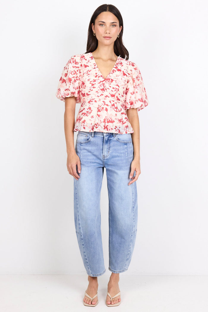 A woman stands against a plain white studio background wearing a short-sleeve floral blouse with puff sleeves, light blue jeans, and clear high-heel sandals.