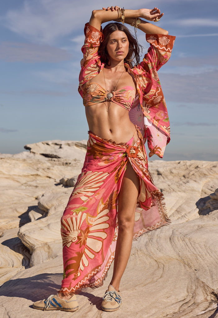 Front view of Oceana Travel Scarf in Coral worn as sarong, showing frayed edge and removable buckle styling on rocks