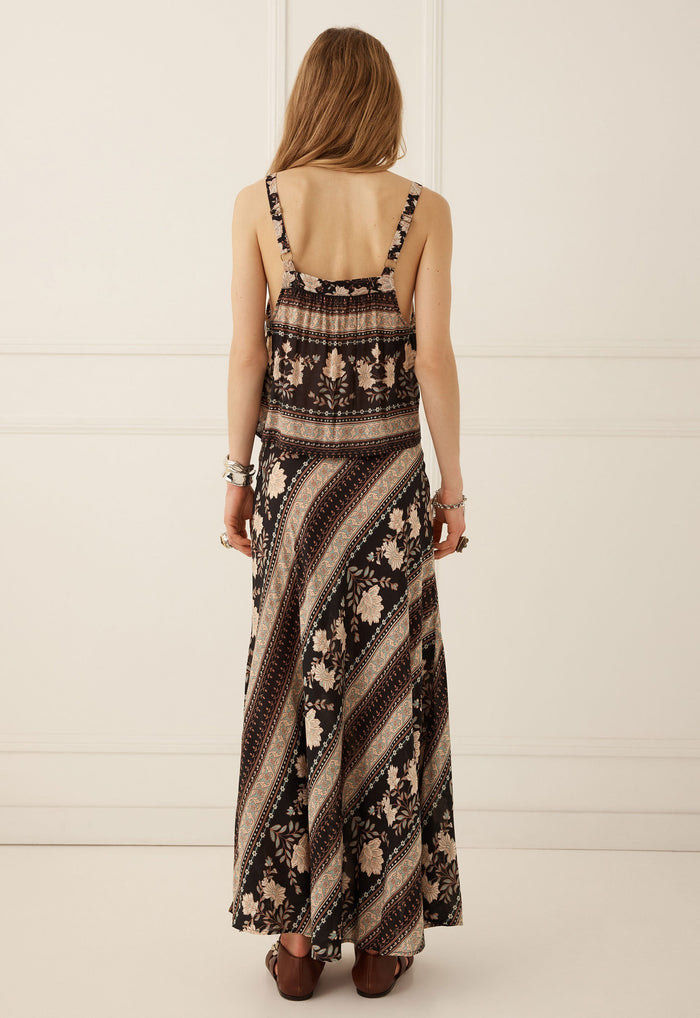 A woman viewed from behind wearing a sleeveless maxi dress with a brown, beige, and black striped and floral pattern, paired with brown flat shoes against a white paneled wall.