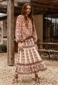 A woman wearing a bohemian-style two-piece dress set with a rust brown and cream floral print, standing on a gravel patio in front of a rustic wooden structure.