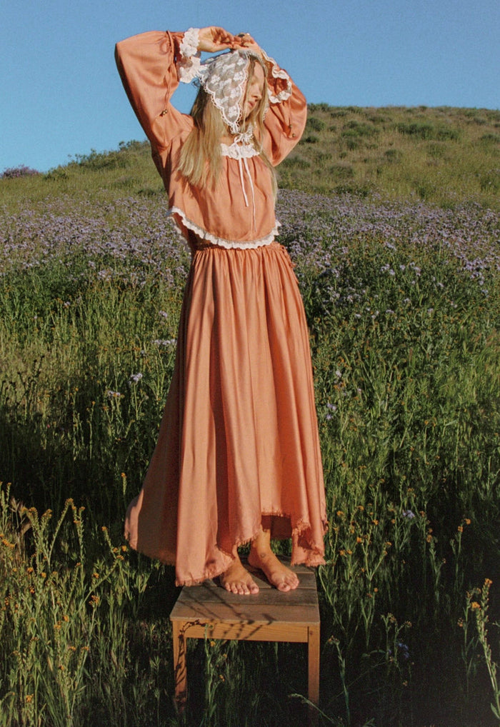 Back view of rose TENCEL blouse on model in wildflower field, showing lace-trim hem and long sleeves with ties
