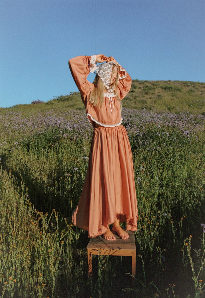 Back view of a model in rose TENCEL maxi skirt, standing on stool in wildflower field, hi-lo frayed hem visible