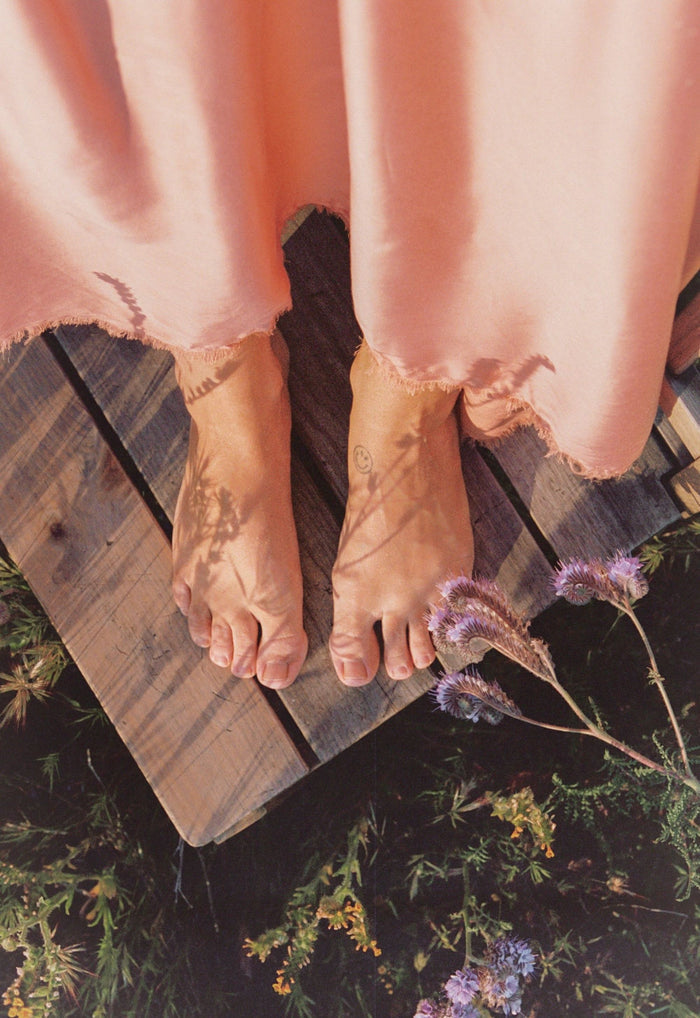 Close-up of frayed TENCEL hem on rose maxi skirt, model barefoot on wooden board among wildflowers