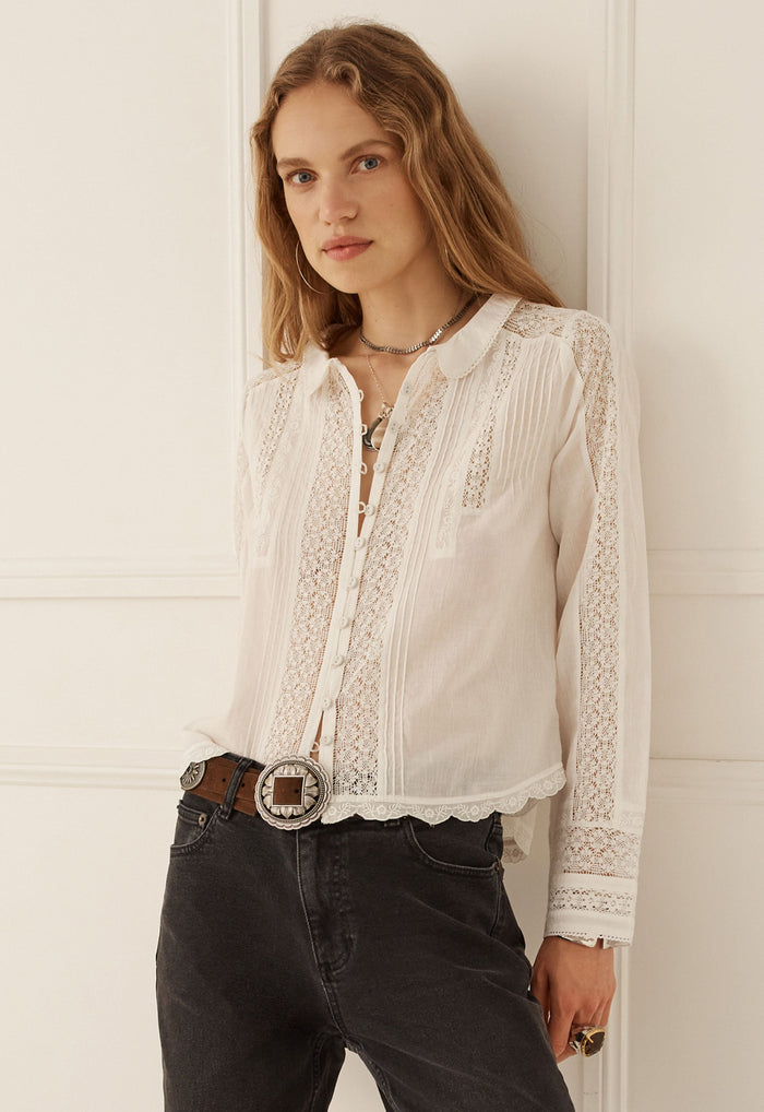A woman wearing a white long-sleeve blouse with intricate lace and embroidered detailing, tucked into dark gray jeans with a brown belt, standing against a white paneled wall.
